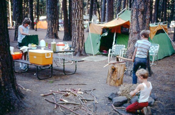 Campsite at Yosemite. Carole fixing dinner on campstove. Tom splitting wood. Bill adding kindling to fire ring. Green tent. Table filled with cooler and foodstuff.