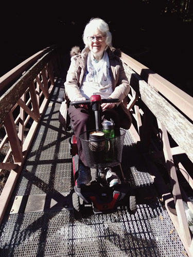 Carole "hiking" in her scooter. She's in the middle of a wooden bridge on a hiking trail.