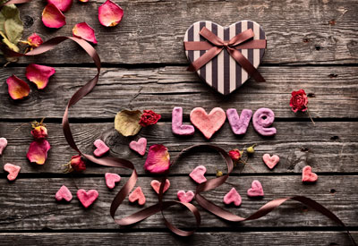 "Love" is spelled out in pink candy hearts on wood background. There is a brown heart-shaped box with a bow. There are heart-shaped candy pieces and flower petals along a curly brown ribbon.