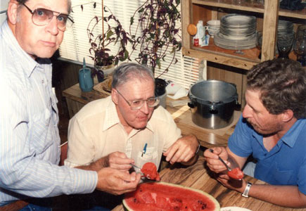 Carole's Dad, sitting, uncle, standing, and cousin, sitting at table. All are enjoying eating from a watermelon half.