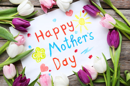 Happy Mothers Day, written in red and blue with hearts and flowers on white sign. Surrounded by pink, purple, and white tulips.